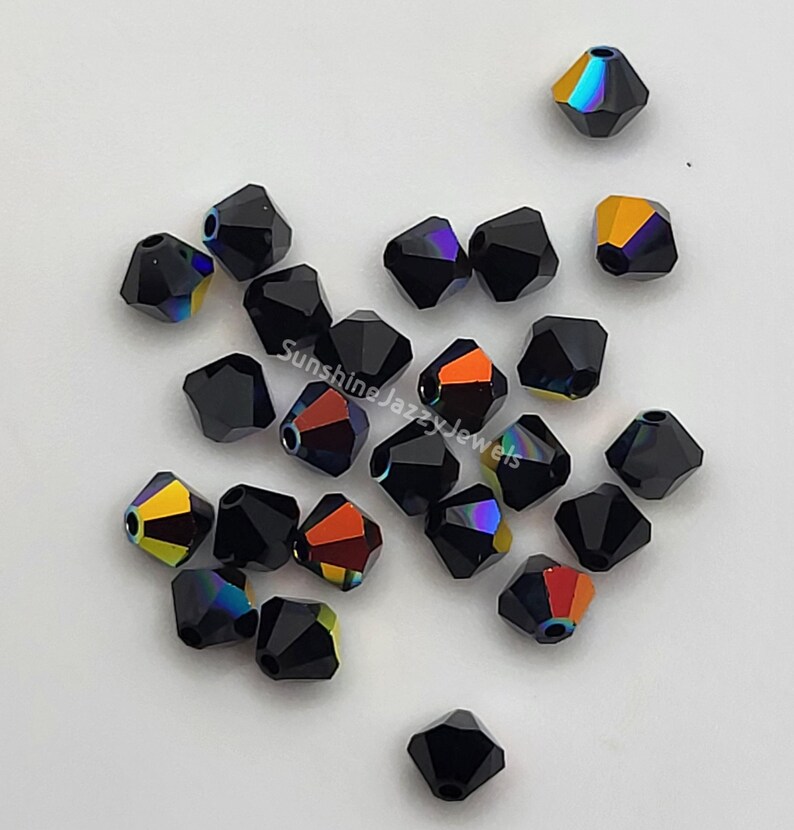 May include: A collection of black, faceted beads with iridescent color flashes of orange, yellow, and purple. The beads are scattered on a white surface. The text "Sunshine Jazzy Jewels" is visible on the image.
