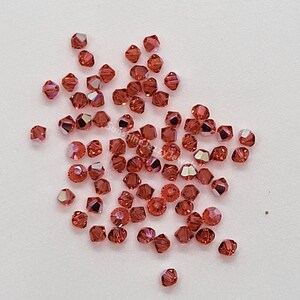 K&ouml;nnte beinhalten: Eine Ansammlung kleiner, facettierter, bikonischer Perlen in einem tiefen Rot-Orange-Ton mit irisierenden Oberfl&auml;chen. Die Perlen sind verstreut und zeigen zahlreiche scharfe Kanten und reflektierende Fl&auml;chen. Der Text SunshineJazzyJewels ist zu sehen.
