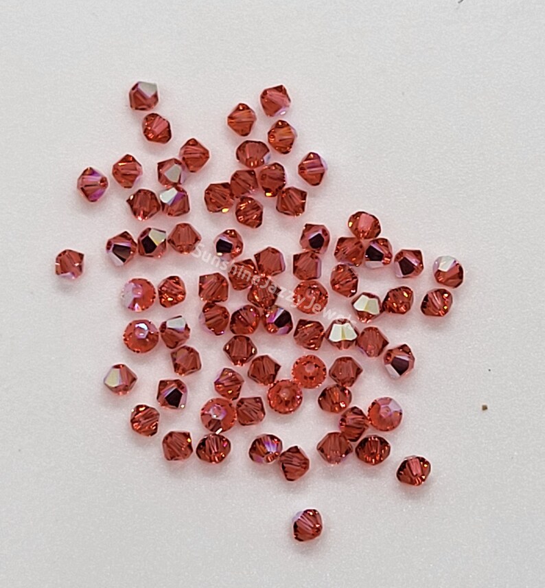 K&ouml;nnte beinhalten: Eine Ansammlung kleiner, facettierter, bikonischer Perlen in einem tiefen Rot-Orange-Ton mit irisierenden Oberfl&auml;chen. Die Perlen sind verstreut und zeigen zahlreiche scharfe Kanten und reflektierende Fl&auml;chen. Der Text SunshineJazzyJewels ist zu sehen.