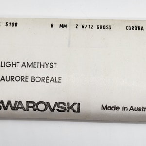 Puede incluir: Una etiqueta blanca con texto negro. Se lee "ART. 5100", "6 MM" y "2 6/12 GROSS". La etiqueta tambi&eacute;n indica "LIGHT AMETHYST AURORE BOR&Eacute;ALE" y "SWAROVSKI Made in Austria".
