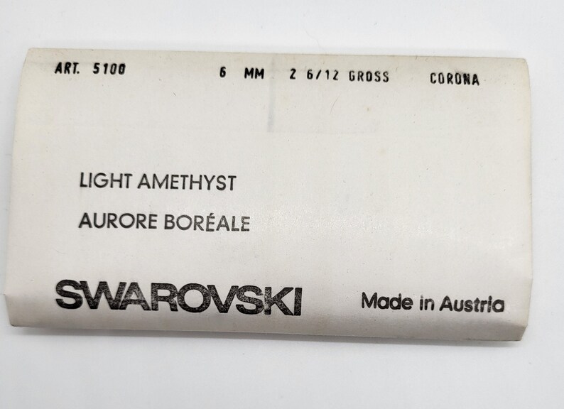 Puede incluir: Una etiqueta blanca con texto negro. Se lee "ART. 5100", "6 MM" y "2 6/12 GROSS". La etiqueta tambi&eacute;n indica "LIGHT AMETHYST AURORE BOR&Eacute;ALE" y "SWAROVSKI Made in Austria".