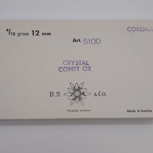 May include: A white box with the text "CORONA" in purple lettering. The box also has the text "Art. 5100", "CRYSTAL COMET OR", "D.S. & Co.", "TRADE MARK", and "Made in Austria". The box is labeled "4/12 gross 12 mm".