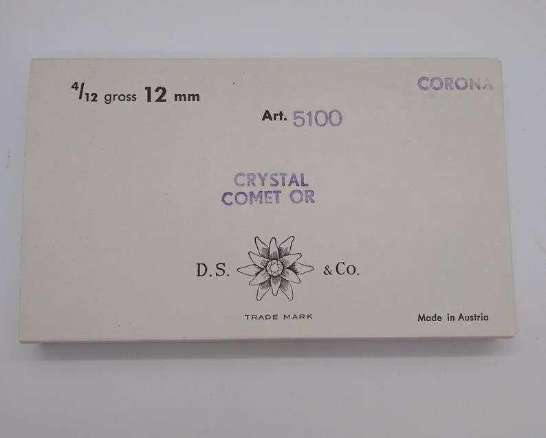 May include: A white box with the text "CORONA" in purple lettering. The box also has the text "Art. 5100", "CRYSTAL COMET OR", "D.S. & Co.", "TRADE MARK", and "Made in Austria". The box is labeled "4/12 gross 12 mm".