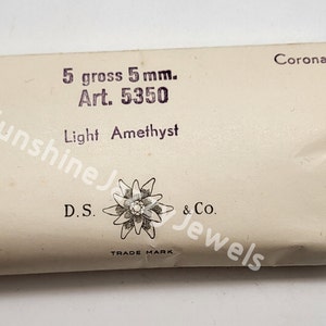 May include: A white package with the text "5 gross 5 mm. Art. 5350 Light Amethyst" and a logo of a stylized flower with the text "D.S. & Co. TRADE MARK" and "Corona" in the top right corner.