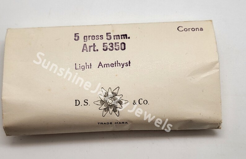 May include: A white package with the text "5 gross 5 mm. Art. 5350 Light Amethyst" and a logo of a stylized flower with the text "D.S. & Co. TRADE MARK" and "Corona" in the top right corner.