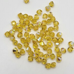 May include: A scattering of faceted, golden-yellow beads with iridescent highlights. The beads are small, with multiple facets that catch the light, creating a sparkling effect. These beads are suitable for jewelry making and craft projects.