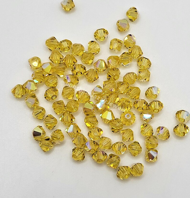 May include: A scattering of faceted, golden-yellow beads with iridescent highlights. The beads are small, with multiple facets that catch the light, creating a sparkling effect. These beads are suitable for jewelry making and craft projects.