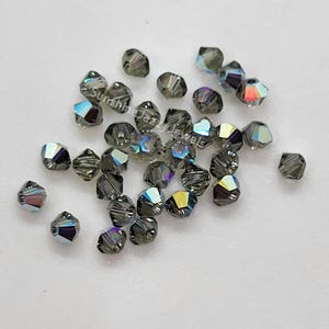 K&ouml;nnte beinhalten: Eine Ansammlung kleiner, facettierter, dunkelgrauer Bikon-Perlen mit irisierendem Finish, das Regenbogenfarben reflektiert. Der Text SunshineJazzyJewels ist &uuml;ber die verstreuten Perlen gedruckt, die eine scharfe, geometrische Form aufweisen.