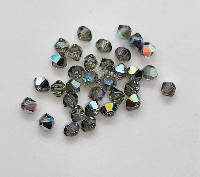 K&ouml;nnte beinhalten: Eine Ansammlung kleiner, facettierter, dunkelgrauer Bikon-Perlen mit irisierendem Finish, das Regenbogenfarben reflektiert. Der Text SunshineJazzyJewels ist &uuml;ber die verstreuten Perlen gedruckt, die eine scharfe, geometrische Form aufweisen.
