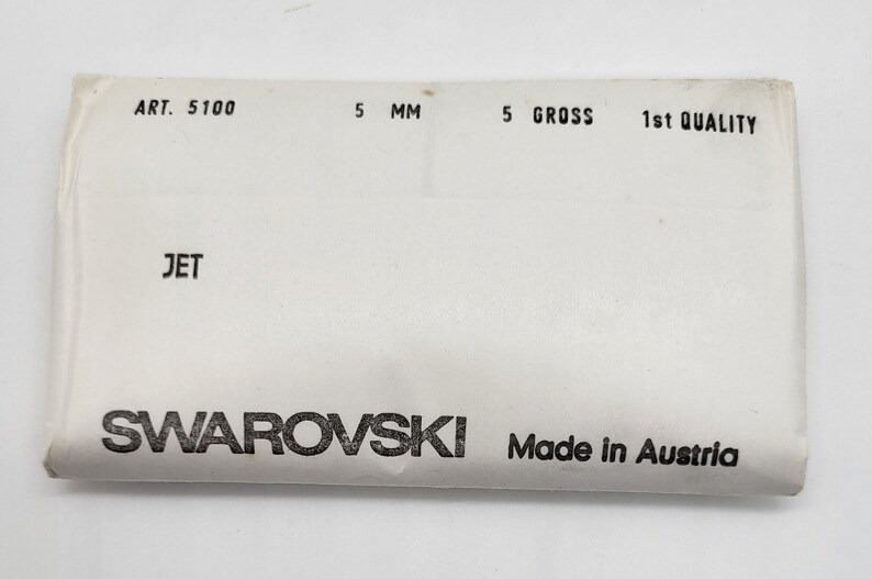 May include: A white package with black text that reads "ART. 5100 5 MM 5 GROSS 1st QUALITY JET SWAROVSKI Made in Austria".