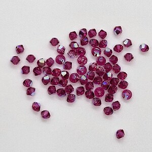 K&ouml;nnte beinhalten: Eine Ansammlung kleiner, facettierter, tief magentafarbener Kristallperlen, die auf einer glatten Oberfl&auml;che verstreut sind. Jede Perle hat eine facettierte Oberfl&auml;che, die das Licht reflektiert und einen schimmernden Effekt erzeugt. Der Text SunshineJazzJewels ist sichtbar.