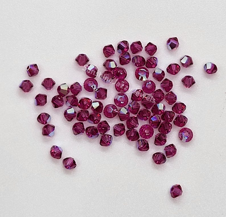 K&ouml;nnte beinhalten: Eine Ansammlung kleiner, facettierter, tief magentafarbener Kristallperlen, die auf einer glatten Oberfl&auml;che verstreut sind. Jede Perle hat eine facettierte Oberfl&auml;che, die das Licht reflektiert und einen schimmernden Effekt erzeugt. Der Text SunshineJazzJewels ist sichtbar.