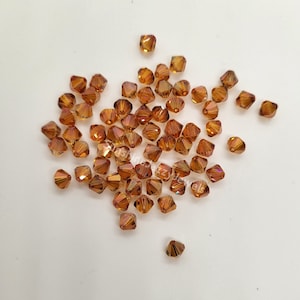 May include: A scattering of amber-colored glass beads with a faceted, pyramid-like shape. The beads are arranged in a loose cluster on a white surface.
