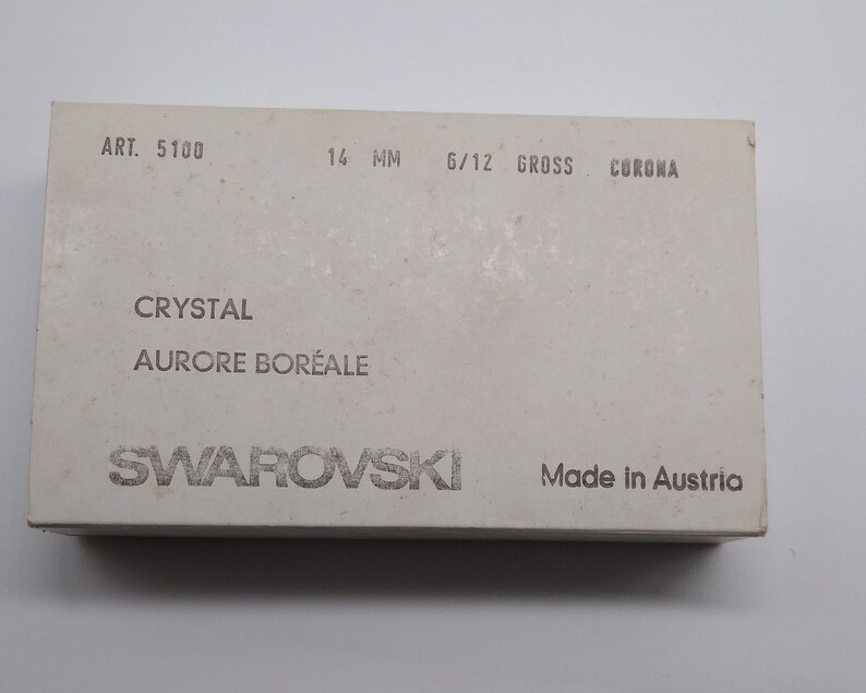 K&ouml;nnte beinhalten: Eine wei&szlig;e Box mit dem Text "ART. 5100 14 MM 6/12 GROSS CORONA" und "CRYSTAL AURORE BOR&Eacute;ALE SWAROVSKI Made in Austria".