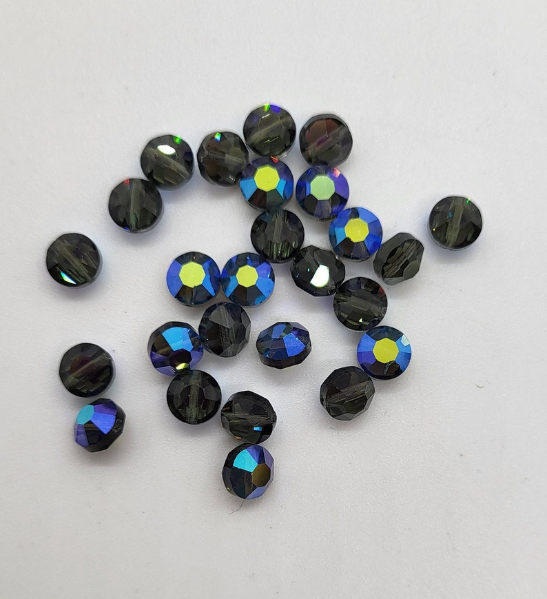 May include: A scattering of dark blue iridescent glass beads, some with a hint of green, on a white background.