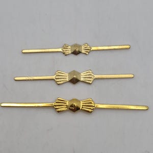 May include: Three gold-toned metal fasteners arranged horizontally. Each piece features a central raised hexagonal shape flanked by two fan-shaped decorative wings, extending into thin, straight bars on both sides of the center.