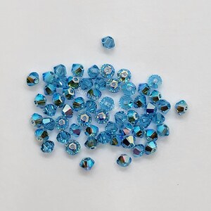 K&ouml;nnte beinhalten: Eine Ansammlung kleiner, facettierter, hellblauer Kristallperlen mit irisierendem Finish. Die Perlen sind bikonisch und scheibenf&ouml;rmig und verf&uuml;gen &uuml;ber ein mittiges Loch zum Auff&auml;deln. Der Text SunshineJazJewelry ist in der Mitte sichtbar.