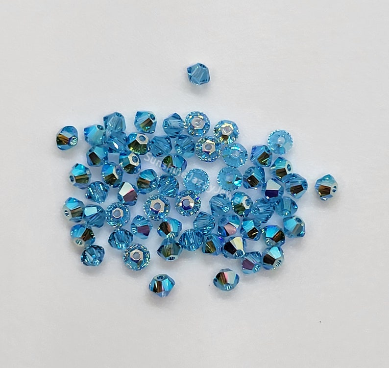 K&ouml;nnte beinhalten: Eine Ansammlung kleiner, facettierter, hellblauer Kristallperlen mit irisierendem Finish. Die Perlen sind bikonisch und scheibenf&ouml;rmig und verf&uuml;gen &uuml;ber ein mittiges Loch zum Auff&auml;deln. Der Text SunshineJazJewelry ist in der Mitte sichtbar.