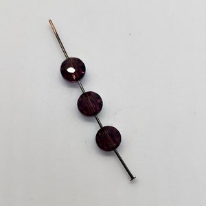 May include: A bead pin featuring three dark purple, round beads. The beads are strung on a thin, dark metal pin with a copper tip. The beads have a faceted surface, reflecting light. The pin is on a white surface.