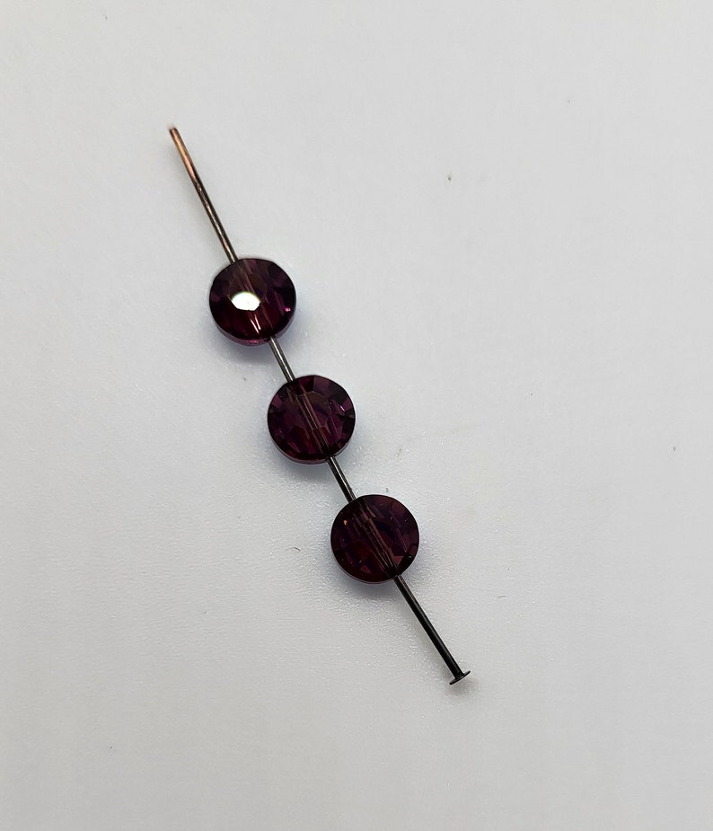 May include: A bead pin featuring three dark purple, round beads. The beads are strung on a thin, dark metal pin with a copper tip. The beads have a faceted surface, reflecting light. The pin is on a white surface.