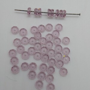 May include: A scattering of light pink glass beads with a faceted design. The beads are arranged in a loose pile on a white surface. A single strand of beads is strung on a wire.