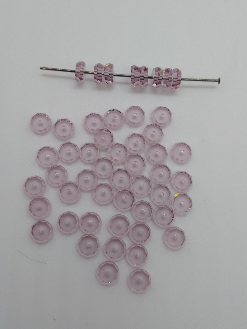 May include: A scattering of light pink glass beads with a faceted design. The beads are arranged in a loose pile on a white surface. A single strand of beads is strung on a wire.