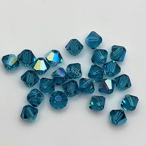 May include: A collection of faceted teal-colored beads. The beads are square-cut with multiple facets that reflect light, creating a shimmering effect. The beads are scattered on a white surface. The text "Sunshine Jazz Jewels" is visible.