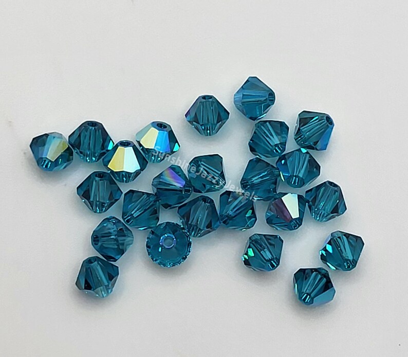May include: A collection of faceted teal-colored beads. The beads are square-cut with multiple facets that reflect light, creating a shimmering effect. The beads are scattered on a white surface. The text "Sunshine Jazz Jewels" is visible.