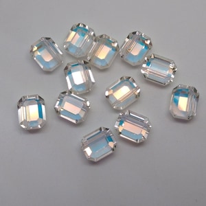 May include: A collection of clear, rectangular crystal beads with iridescent, rainbow-like reflections. The beads are faceted, catching and reflecting light. These beads are suitable for jewelry making and crafting.