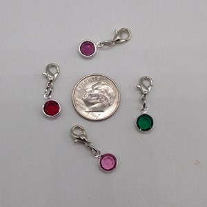 May include: Four silver charm clips with different colored rhinestones. The charms are pink, red, green, and purple. The charms are about the size of a dime.