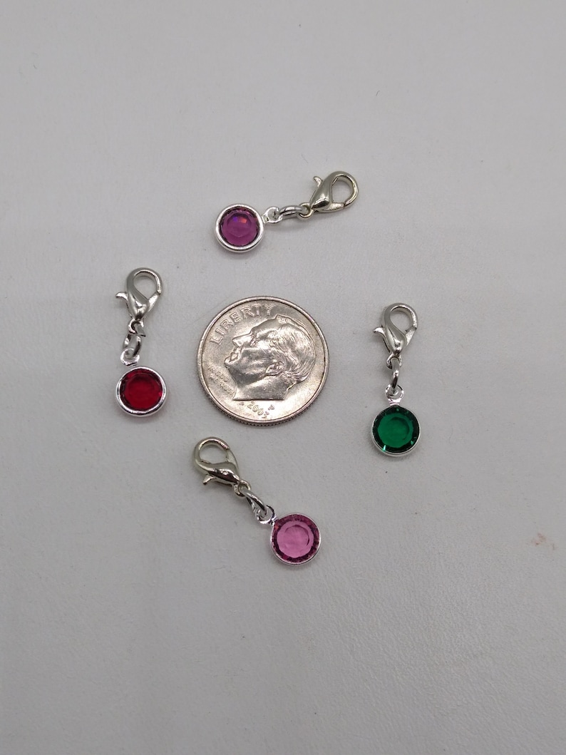 May include: Four silver charm clips with different colored rhinestones. The charms are pink, red, green, and purple. The charms are about the size of a dime.