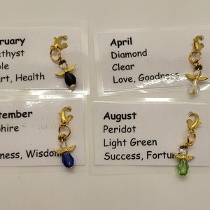 May include: Four small cards with gold charms attached. Each card features a month of the year, the corresponding birthstone, its color, and associated qualities. The charms are angel-shaped with a crystal bead in the center. The crystals are black, clear, blue, and light green.