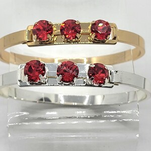 May include: Two bracelets with three red gemstones each. The top bracelet is gold and the bottom bracelet is silver.