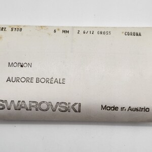 May include: A white card with black text that reads "ART. 5100 6 MM 2 6/12 GROSS CORONA MORION AURORE BOR&Eacute;ALE SWAROVSKI Made ir. Austria"