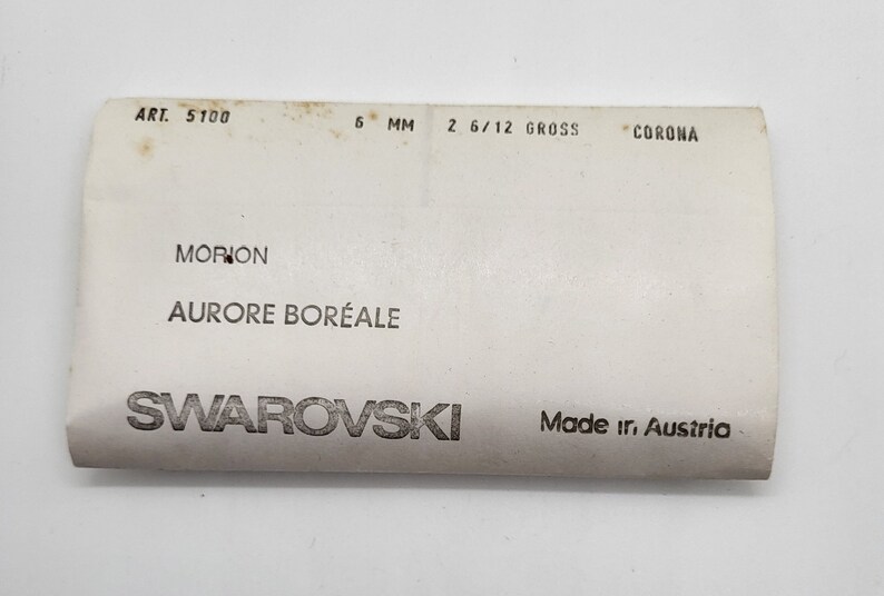 May include: A white card with black text that reads "ART. 5100 6 MM 2 6/12 GROSS CORONA MORION AURORE BOR&Eacute;ALE SWAROVSKI Made ir. Austria"