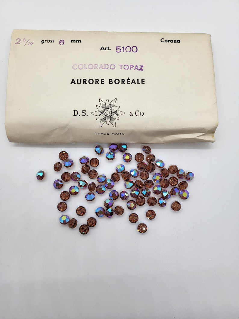 May include: A small package of 6 mm brown glass beads with iridescent rainbow coloring. The package is labeled "Corona Art. 5100 COLORADO TOPAZ AURORE BOR&Eacute;ALE D.S. & Co. TRADE MARK"