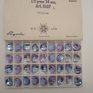 May include: A box of iridescent, faceted beads, each approximately 0.55 inches in diameter, arranged in a grid. The beads have a mix of purple, blue, and silver tones. The box is labeled with text including "Wistaria" and "Made in Austria."