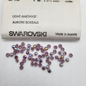 Puede incluir: Cristales Swarovski amatista claro, de 6 mm de di&aacute;metro, con efecto aurora boreal. Los cristales est&aacute;n dispersos sobre una superficie blanca junto a una etiqueta que dice "Light Amethyst, Aurore Bor&eacute;ale, Swarovski, Made in Austria."