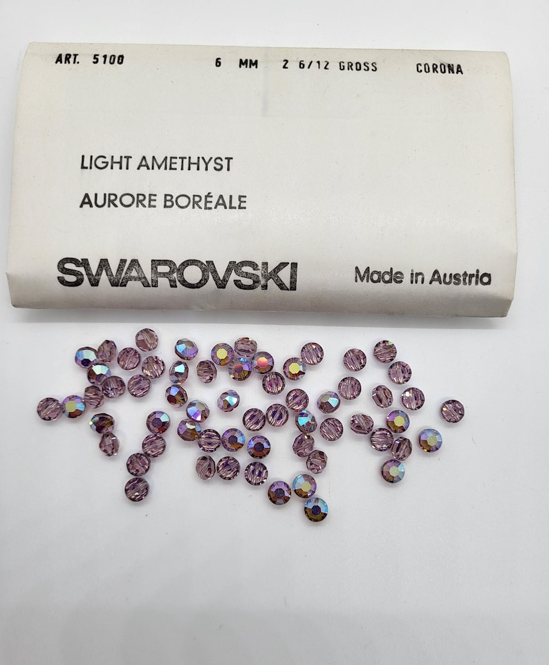 Puede incluir: Cristales Swarovski amatista claro, de 6 mm de di&aacute;metro, con efecto aurora boreal. Los cristales est&aacute;n dispersos sobre una superficie blanca junto a una etiqueta que dice "Light Amethyst, Aurore Bor&eacute;ale, Swarovski, Made in Austria."