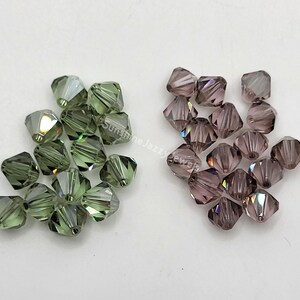 May include: Two piles of faceted beads. One pile is olive green, the other is a muted purple. The beads are square-cut with multiple facets, reflecting light. The beads are likely for jewelry making.