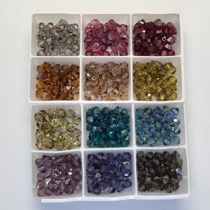 May include: Assortment of 12 small glass beads in various colors, including clear, pink, red, brown, gold, green, blue, purple, and gray. The beads are arranged in a white plastic tray with 12 compartments.