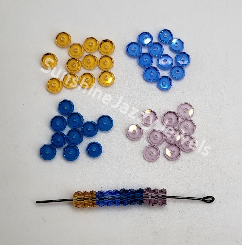May include: Assortment of small, faceted glass beads in four colors: gold, blue, light blue, and lavender. The beads are arranged in four piles on a white surface. A single strand of beads in gold, blue, and lavender is shown in the bottom center of the image.