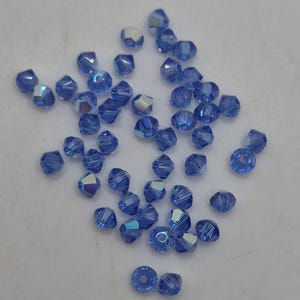 K&ouml;nnte beinhalten: Eine Ansammlung kleiner, facettierter Bikon-Perlen in leuchtendem Kornblumenblau. Jede Perle besitzt eine reflektierende, irisierende Oberfl&auml;che, die das Licht einf&auml;ngt und subtile Glanzlichter auf den vielen kantigen Fl&auml;chen des Glases erzeugt.