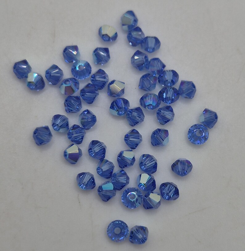 K&ouml;nnte beinhalten: Eine Ansammlung kleiner, facettierter Bikon-Perlen in leuchtendem Kornblumenblau. Jede Perle besitzt eine reflektierende, irisierende Oberfl&auml;che, die das Licht einf&auml;ngt und subtile Glanzlichter auf den vielen kantigen Fl&auml;chen des Glases erzeugt.