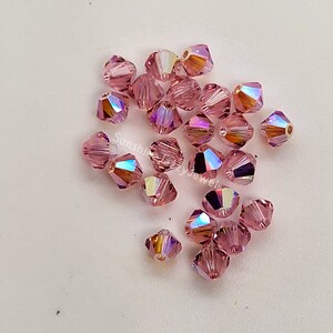 May include: A collection of faceted, light pink crystal beads. Each bead has multiple facets that reflect light, creating a rainbow effect. The beads are scattered on a white surface. The text "Sunshine Crazy Jewels" is visible.