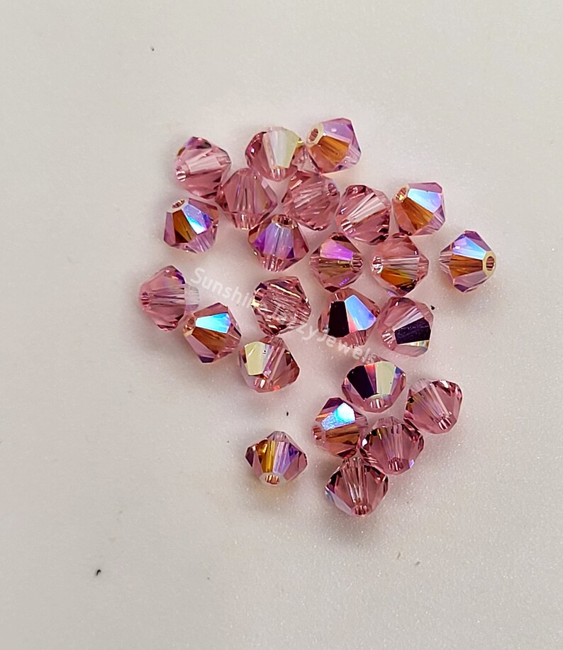May include: A collection of faceted, light pink crystal beads. Each bead has multiple facets that reflect light, creating a rainbow effect. The beads are scattered on a white surface. The text "Sunshine Crazy Jewels" is visible.