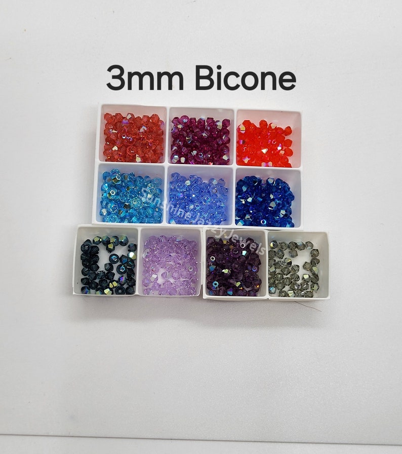 K&ouml;nnte beinhalten: Ein wei&szlig;es Kunststoff-Sortierfach mit neun F&auml;chern, gef&uuml;llt mit 3 mm Bicone-Perlen in verschiedenen Farben wie Rot, Magenta, Orange, Hellblau, Mittelblau, Dunkelblau, Schwarz, Lavendel, Violett und Grau mit dem Text 3mm Bicone.