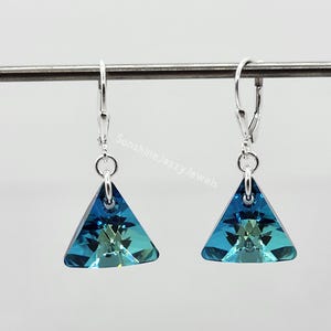 May include: A pair of leverback earrings with triangular, iridescent blue and green crystal pendants. The earrings are suspended from silver-colored metal hooks. The image is set against a plain background.