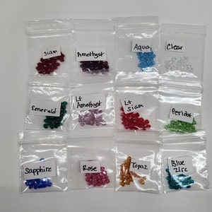 Könnte beinhalten: Zwölf kleine Plastiktüten, die jeweils eine andere Farbe von kleinen, facettierten Glasperlen enthalten. Die Beutel sind mit der Farbe der Perlen im Inneren beschriftet: Siam, Amethyst, Aqua, Klar, Smaragd, Hell-Amethyst, Hell-Siam, Peridot, Saphir, Rose, Topas und Blauer Zirkon.