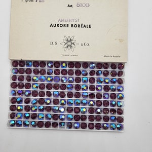May include: A box of amethyst-coloured, iridescent rhinestones, each 7 mm in diameter, arranged in a grid. The package includes text that reads "AMETHYST AURORE BOR&Eacute;ALE" and "Made in Austria."